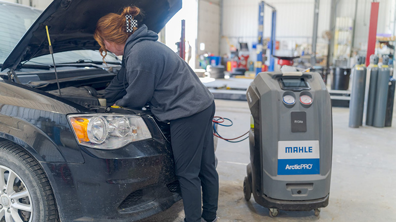 Red-haired technician leaning under the hood of a dark minivan connected to an A/C recharge machine
