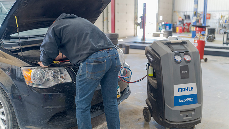 Mechanic working under the hood of a car hooked up to an A/C recharge machine in an auto repair shop