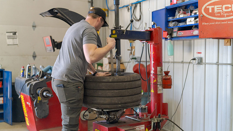 Technician mounting a tire on a red tire changer in an auto shop, with a wheel balancer behind him