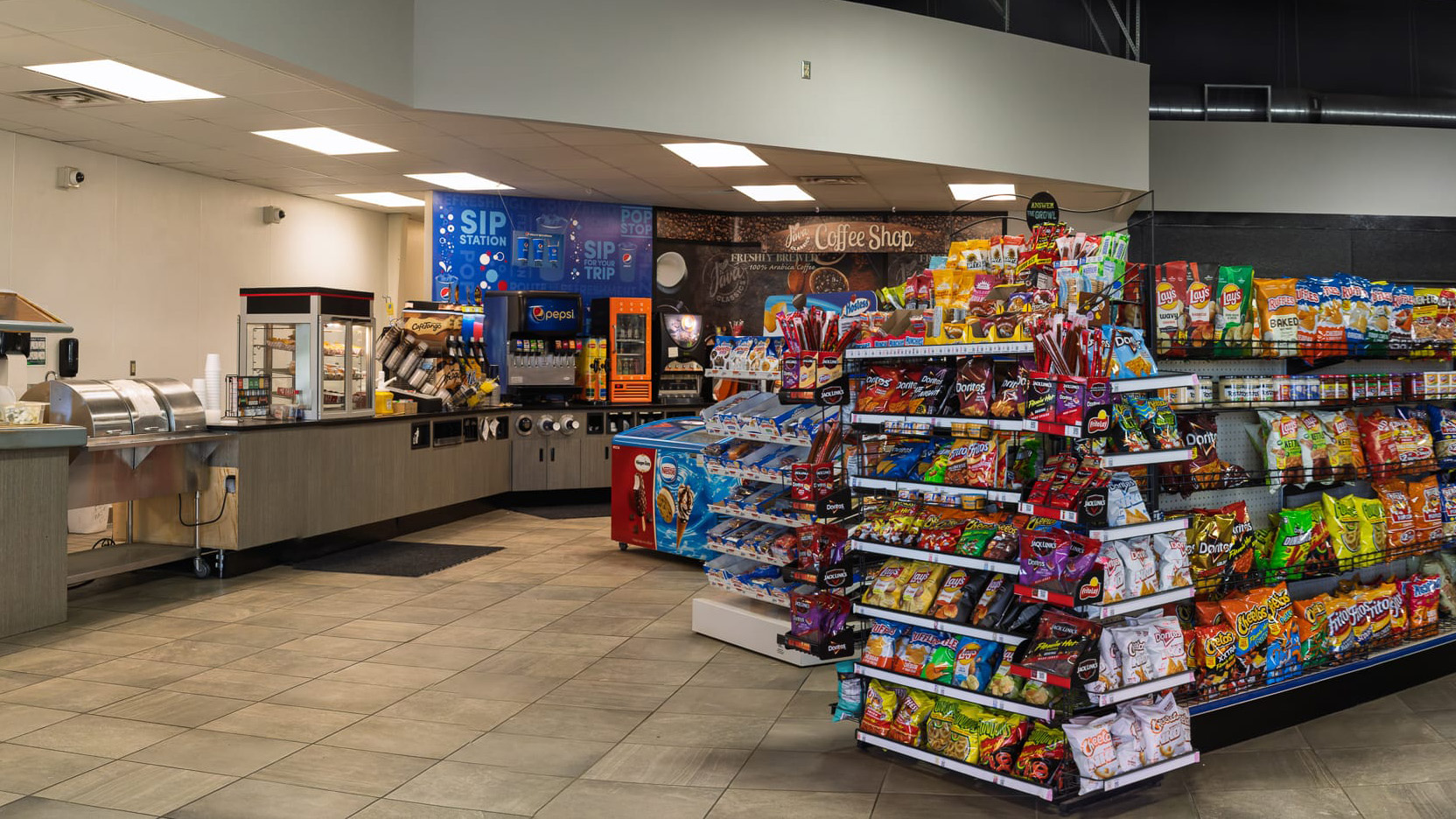 Convenience store interior with a hot food counter, fountain drinks, coffee station, and aisles of chips and snacks