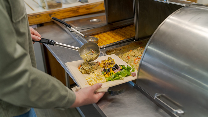 Person ladling a rice casserole onto a plate of mac and cheese and salad at a stainless steel hot food bar