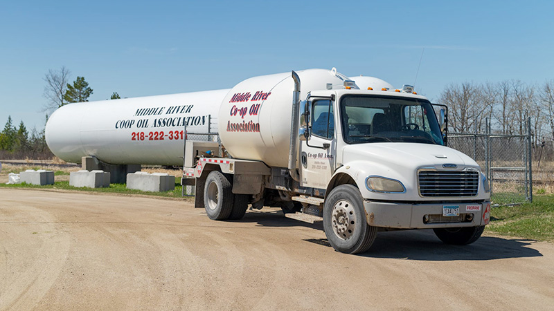 Front view of a Middle River Co-op Oil propane delivery truck on a gravel lot, with the bulk storage tank behind it